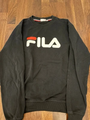 Svart sweatshirt från FILA - Snygg svart sweatshirt från FILA med stort vitt och rött logotryck på bröstet. Tröjan har rund halsringning, ribbade muddar vid ärmslut och nederkant samt en avslappnad passform. OBS! Den är stor för sin storlek 