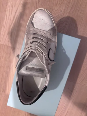 Vita sneakers från Philippe Model storlek 43 - Snygga vita sneakers från Philippe Model med klassisk låg profil, svarta detaljer vid hälen och den ikoniska sköld-loggan på sidan. Skorna är gjorda i läder med mockadetaljer och har vita snören. Perfekta för en trendig streetstyle-look. Dock lite söndriga på undersidan men är väldigt enklet o smidigt att laga. Priset kan diskuteras vid snabb affär❗️