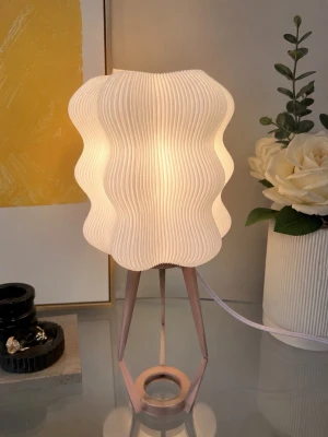 Modern bordslampa med vågig skärm - Säljer en modern bordslampa med vit, vågformad lampskärm i veckad plast och ben i ljusrosa metall. Lampan har en unik design som ger ett mjukt och behagligt ljus, perfekt som inredningsdetalj i sovrum eller vardagsrum.