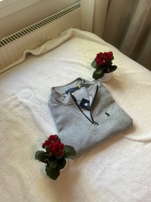 Grå halvzip tröja från Polo Ralph Lauren - Snygg grå sweatshirt med halv dragkedja från Polo Ralph Lauren. Tröjan har en klassisk passform, broderad logga på bröstet och ribbade muddar. Perfekt till jeans eller chinos för en clean och stilren look.