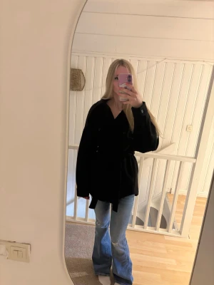 Svart oversized medellång kappa - Snygg svart medellång kappa med klassisk krage och knäppning framtill. Modellen är rak och lite oversized, med två bröstfickor och bälte i midjan för en cool look. Perfekt att slänga över en t-shirt eller hoodie.