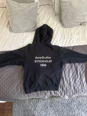 Svart hoodie Acne Studios Stockholm - Svart hoodie från Acne Studios med tryck i vitt på ryggen: 'Acne Studios STOCKHOLM 1996'. Klassisk passform med huva och ribbade muddar. Perfekt för en stilren och avslappnad look. Materialet känns mjukt och skönt mot huden.