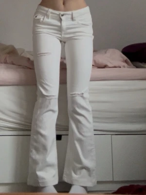 Vita bootcut jeans med slitningar - Säljer ett par vita bootcut jeans med slitningar vid knäna och låret. Jeansen har låg midja, klassiska fem fickor och bakfickor med lock och knapp. Perfekta för en trendig och avslappnad stil. Materialet är denim i bomull.