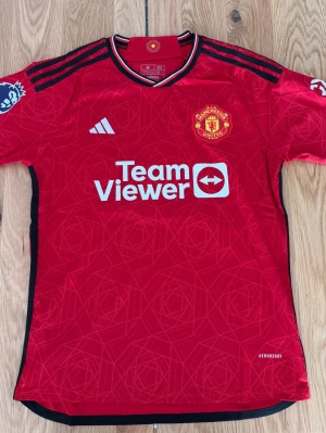 Manchester United matchtröja Adidas - Röd Manchester United matchtröja från Adidas med svarta detaljer och klubbmärke på bröstet. Tröjan har TeamViewer-logga framtill, Premier League-märke på ärmen och är tillverkad i lätt funktionsmaterial (Aeroready). Perfekt för fotbollsträning eller support.
