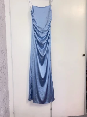 Ljusblå långklänning i satin - Elegant ljusblå långklänning i satin med tunna axelband och draperad framsida. Klänningen har en öppen rygg och en slank, följsam passform som ger en stilren och modern look. Perfekt för speciella tillfällen.