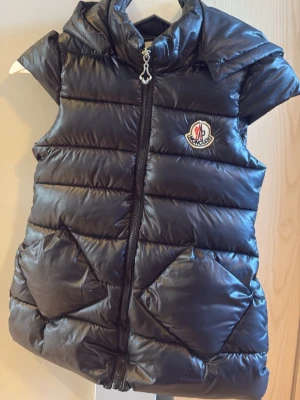 Svart dunväst från Moncler - Snygg svart dunväst från Moncler med glansig finish, dragkedja framtill och ikonisk logotyp på bröstet. Västen har hög krage, två fickor och quiltad design som ger en cool och sportig look.