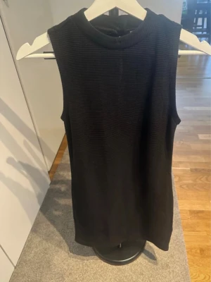 Svart ribbad ärmlös kort klänning  - Snygg svart ärmlös kort klänning med ribbad struktur ocht hög hals. Klänningen har en enkel och stilren design med knäppning i nacken. 