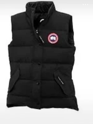 Svart dunväst från Canada Goose - Snygg svart dunväst från Canada Goose med klassisk logga på bröstet. Västen har hög krage, dragkedja och knappar framtill samt två fickor med lock. Perfekt för lager-på-lager och håller dig varm under kyliga dagar. Storlek xs men passar även S 