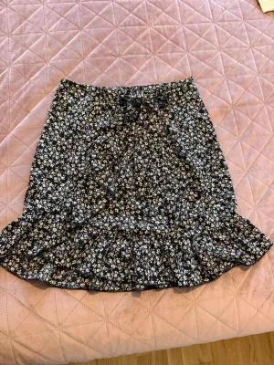 Svart blommig volangkjol - Snygg svart kjol med vitt blommönster och volangkant nertill. Kjolen har en rak passform upptill och en bred volang som ger en trendig look. Perfekt för dig som gillar feminina detaljer och mönster. Materialet känns lätt och luftigt.