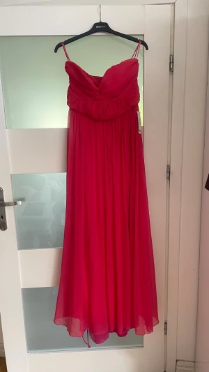 GINA TRICOT Röd rosa långklänning  - Säljer en elegant röd långklänning, draperad överdel. Klänningen har ett flytande fall och är tillverkad i ett lätt, skirt material som ger en romantisk känsla. Perfekt för speciella tillfällen.