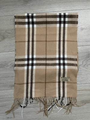 Beige rutig halsduk från Burberry - Klassisk beige halsduk från Burberry i 100% lammull med det ikoniska rutmönstret i brunt, svart och vitt. Halsduken har fransar i ändarna och är tillverkad i England. Perfekt accessoar för att lyfta din outfit.