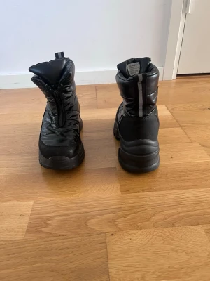 Svarta moon boots med dragkedja - Säljer ett par svarta moon boots med chunky sula och dragkedja framtill. Skorna är vadderade och har en glansig finish i syntetmaterial, perfekta för kalla dagar. De har en modern och sportig look som passar till vinterns outfits. Storlek35