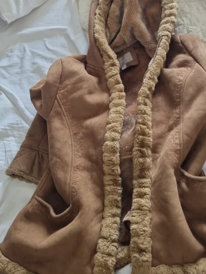 Brun fuskpälsfodrad kappa med huva - Mysig brun kappa med fuskpälsfoder och huva. Kappan har dekorativa sömmar, två fickor och fluffiga kanter längs öppningen och huvan. Perfekt för kalla dagar när du vill vara både varm och snygg.