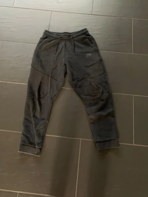 Svarta Nike tech mjukisbyxor med dragsko - Svarta mjukisbyxor från Nike med dragsko i midjan och resår vid bensluten. Byxorna har en sportig design med synliga sömmar och en liten Nike-logga på ena benet. Perfekta för chill eller träning, tillverkade i mjukt material.