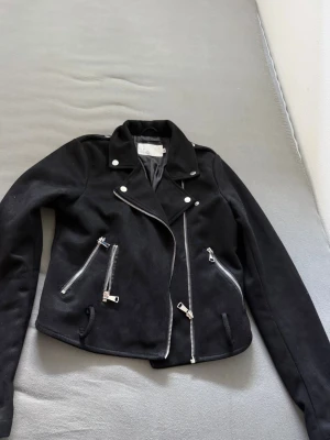 Svart skinnjacka med dragkedjor - Snygg svart skinnjacka i bikerstil med silvriga dragkedjor och detaljer. Jackan har snedställd dragkedja framtill, två fickor med dragkedja och dekorativa knappar vid kragen. Kortare modell som ger en cool look.