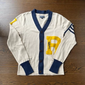Cardigan från Ralph Lauren  - Beige cardigan med blåa muddar och knappar från Polo Jeans Company (Ralph Lauren) . Stora gula bokstaven R och siffran 67 på framsidan, samt blåa och gula detaljer på ärmarna. Klassisk collegestil med v-ringning och ribbade muddar. PRIS KAN DISKUTERAS