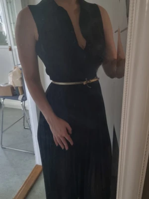 Svart långklänning med bälte från Calvin Klein - Svart ärmlös långklänning med djup v-ringning och ett smalt ljust bälte i midjan. Klänningen har en elegant och tidlös design, perfekt för dig som gillar stilrena plagg. Materialet känns lätt och följsamt. Hållaren för bältet på ena sidan är trasig (se bild). 