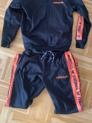 Svart och orange träningsset Askari - Säljer ett svart träningsset från Askari med orangea detaljer och logga på både jacka och byxor. Setet har dragkedja på jackan, resår och snörning i midjan samt orangea ränder med Askari-tryck längs ärmar och ben. Perfekt för gym eller sport.