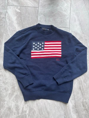  Polo Ralph Lauren - Säljer en marinblå stickad tröja från Polo Ralph Lauren med amerikansk flagga i rött, vitt och blått på bröstet. Tröjan har rund halsringning, ribbade muddar och RL-broderi. Perfekt för dig som gillar klassisk och preppy stil.