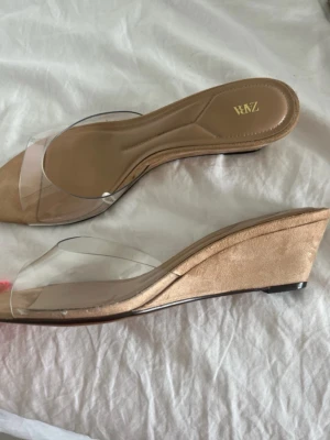 Beige kilklackade sandaler från Zara - Säljer ett par snygga sandaler från Zara med beige mockaliknande sula och transparent rem över foten. Skorna har en låg kilklack och öppen tå, vilket ger en modern och stilren look. Perfekta för att lyfta din outfit med en trendig touch. Är köpta på Zara men i fel storlek, men behövde ett par så fick bli dessa för en användning därav säljs de