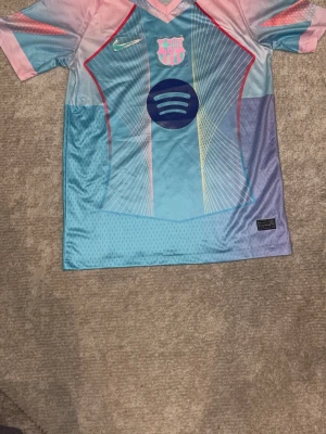 FC Barcelona Nike matchtröja blå/rosa - Snygg FC Barcelona matchtröja från Nike i blått och rosa med klubbmärke och Spotify-logga på bröstet. Tröjan har korta ärmar, sportig passform och är tillverkad i lätt, ventilerande material. Perfekt för fotbollsträning eller att bära som supporter.