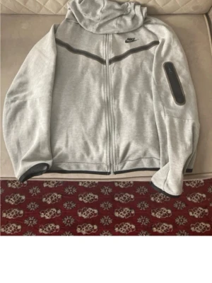 Ljusgrå Nike hoodie med dragkedja - Snygg ljusgrå hoodie från Nike med hel dragkedja framtill och svart detalj längs bröstet. Huva och en praktisk ficka med dragkedja på ärmen. Perfekt för en sportig och avslappnad stil.