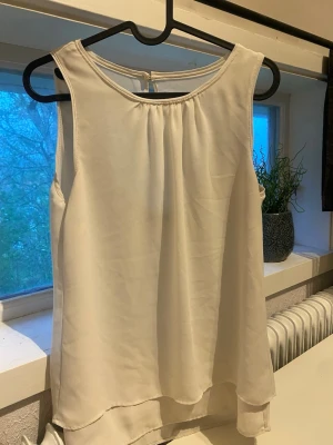 Vit ärmlös linnetopp - Stilren vit ärmlös linnetopp med rund halsringning och lätt transparent material. Toppen har en lös passform och ett diskret fall baktill som ger en snygg siluett. Perfekt att matcha med jeans eller kjol för en fräsch look.