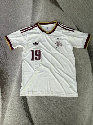 Spanien fotbollströja Adidas #19 - Riktigt fet vit Spanien fotbollströja från Adidas med vinröda detaljer, nummer 19 och Lamine Yamal på ryggen. Tröjan har korta ärmar, rund hals med gul och vinröd kant samt tre klassiska Adidas-ränder på axlarna. Tillverkad i lätt och ventilerande material. Passar perfekt inför sommarn! Priset är inte skrivet i sten.
