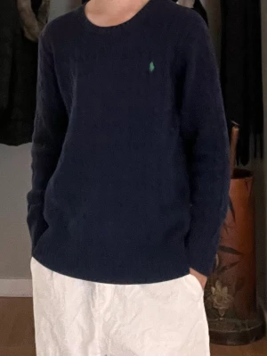 Mörkblå kabelstickad tröja Polo Ralph Lauren - Mörkblå kabelstickad tröja från Polo Ralph Lauren med rund halsringning och grönt broderat logomärke på bröstet. Tröjan har långa ärmar och ribbade muddar vid ärmslut och nederkant. Perfekt för en stilren och klassisk look.