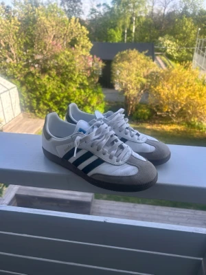 Adidas Samba OG vita och grå sneakers - Adidas Samba OG sneakers i vitt läder med grå mockadetaljer och svarta ränder på sidorna. Klassisk låg modell med svart sula och blå logga på plösen. Perfekta för en stilren och sportig look. Använd endast 3 gånger då jag märkte att de var lite för små för mig, öppen för pris förslag 