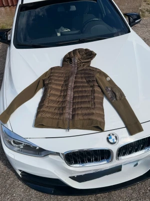 Olivgrön pufferjacka med huva från Moncler - Snygg Omivgrön pufferjacka från Moncler med stickade ärmar och huva. Jackan har dragkedja framtill, ribbade muddar och en ficka med dragkedja på ärmen. Perfekt för kalla dagar och riktigt trendig look.Nypris 6000-7000 mitt pris 2699!!Använd endast 3 gånger!