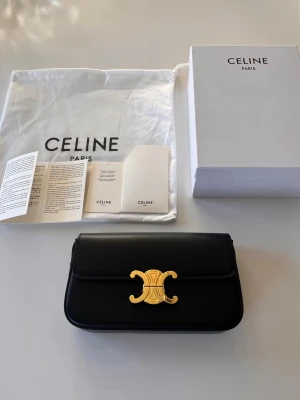 Svart Triomphe handväska från Celine - Säljer en svart handväska från Celine med ikoniskt Triomphe-spänne i guld. Väskan är tillverkad i slätt läder och har en stilren, rektangulär form med lock och axelrem. Kommer med original dustbag, box och äkthetskort. Perfekt för dig som gillar lyxiga detaljer.