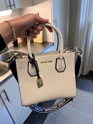 Vit handväska från Michael Kors - Snygg vit handväska från Michael Kors i strukturerat läder med mörkblåa detaljer och guldfärgade metalldelar. Väskan har två handtag, avtagbar axelrem med logga och dekorativt hänglås framtill. Perfekt för dig som gillar stilrena accessoarer. Köpt på mall of Scandinavia i konfa present men knappt använt💕köpt för 5500kr💕