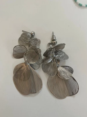 örhängen - Stilrena örhängen i silverfärgad metall med flera lager av tunna, blomformade plattor som ger ett unikt och trendigt intryck. Perfekta för att lyfta din look med en modern touch. Örhängena har stift och är lätta att bära.