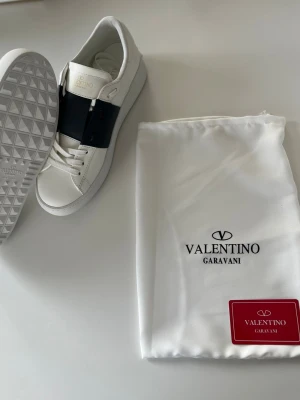 Vita Valentino Garavani sneakers - Snygga vita sneakers från Valentino Garavani med svart band över vristen och diskret logga på plösen. Skorna har en klassisk låg siluett och är tillverkade i läder med vita snören och detaljer. Perfekta för dig som gillar stilrena och exklusiva sneakers.