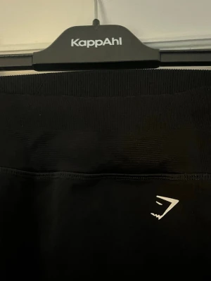 Svarta seamless leggings Gymshark - Svarta träningstights från Gymshark med hög midja och diskret logga framtill. Tightsen har ribbade detaljer/snygga hål längs benen för extra stil och komfort. Perfekta för gymmet eller löprundan. I storlek XS❣️