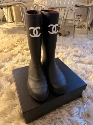 Svarta gummistövlar från Chanel - Snygga svarta gummistövlar från Chanel med det klassiska vita CC-logotypen framtill. Stövlarna är höga och har en stilren, minimalistisk design som passar perfekt till regniga dagar. Tillverkade i slitstarkt gummi för att hålla fötterna torra och bekväma. Lådan är trasig, vilket syns på priset ❤️