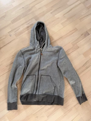 Grå zip-hoodie från L.O.G.G. - En grå hoodie med dragkedja från L.O.G.G. med två stora fickor framtill och kontrastfärgade muddar. Huvan har dragsko och insidan är mjuk och skön. Perfekt för en avslappnad stil.