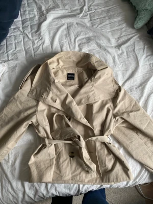 Beige trenchcoat från Bershka - Stilren beige trenchcoat från Bershka med klassisk krage, knytskärp i midjan och axelklaffar. Jackan har dubbelknäppning framtill och är tillverkad i ett lätt bomullstyg. Perfekt för dig som vill ha en tidlös och trendig look.