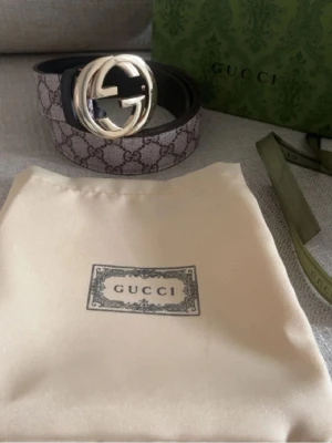  GG Supreme bälte med guldspänne - Snyggt GG Supreme bälte med klassiskt monogrammönster och stort, rundat guldspänne. Bältet har beige och bruna toner med det ikoniska -mönstret och är tillverkat i skinn och canvas. Perfekt accessoar för att lyfta din outfit.