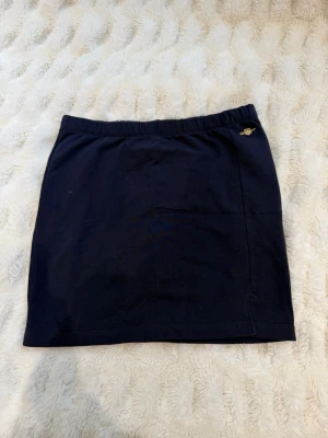 Mörkblå kortkjol från Gant - Mörkblå kortkjol från Gant med elastisk midja och liten guldfärgad logga framtill. Kjolen är enkel och stilren, perfekt att matcha med en t-shirt eller hoodie. Mjuk bomullskvalitet för en bekväm känsla.
