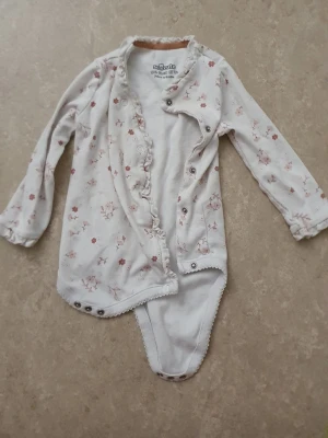 Babybody - Supersöt långärmad body från Newbie i mjuk vit bomull med små blommor i rosa och beige toner. Bodyn har volangkant längs knäppningen framtill och tryckknappar i grenen och på sidan för enkel påklädning. Perfekt för dig som gillar romantiska detaljer.