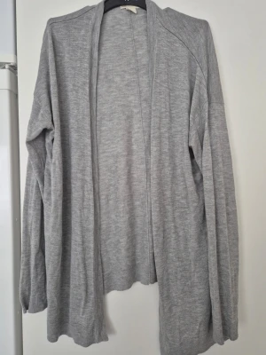 Kofta - Mysig och stilren ljusgrå kofta med lång passform och öppet fall framtill. Tillverkad i mjukt, tunt stickat material som ger en avslappnad look. Perfekt att slänga över en t-shirt eller topp för extra lager.