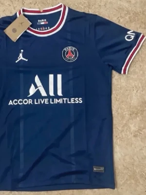 PSG matchtröja blå Jordan - Snygg blå Paris Saint-Germain matchtröja från Jordan med vita och röda detaljer på krage och ärmslut. PSG-logga och Jordan-logga på bröstet samt sponsortryck framtill. Perfekt för fotbollsträning eller att bära som supporter.