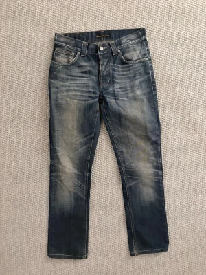 Nudie jeans - Säljer ett par blå jeans från Nudie Jeans med snyggt slitna detaljer och klassisk femficksmodell. Jeansen har raka ben och tydliga kontrastsömmar. Tillverkade i denim med en cool tvätt och Nudie-logga på bakfickan. Size 28:32. 