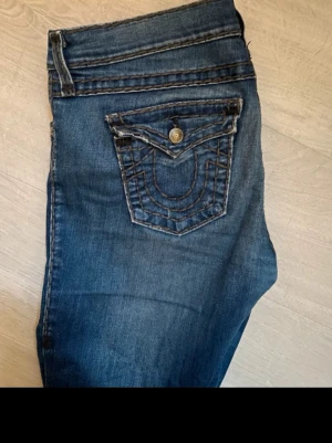 Blå jeans från True Religion - Säljer ett par blå jeans från True Religion med ikonisk kontrastsöm och detaljerad bakficka med lock och knapp. Perfekta till en avslappnad och trendig look. De är i storlek 28. De har lite slitningar vid hälarna och mellan benen (se bilder) men inget som stör 😊