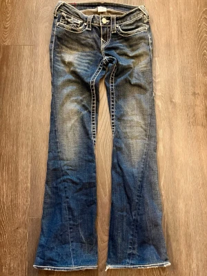 True Religion strl 26, bootcut jeans blå - Säljer ett par blå bootcut jeans från True Religion Strl: 26, med vita kontrastsömmar och slitna detaljer vid benslut.                                            Jeansen har klassiska fickor med lock och emblem bak samt en låg midja. Passformen är utsvängd. Modellen är Limited edition och svår att få tag i,  storleken är något mindre än vanligt. Det sitter strass-stenar på byxorna, men någon har trillat bort (se bild). Den pyssliga kan lätt klistra dit en  ny skulle jag tro!