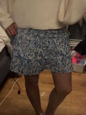 Blåvit blommig kortkjol med volang - Supersöt kortkjol i blått och vitt med blommigt mönster och volangdetaljer. Kjolen har resår i midjan och är perfekt för varmare dagar. Lätt och luftig känsla, passar till många olika toppar. Storlek 146-152 men passar mig helt okej och då har jag vanligtvis s