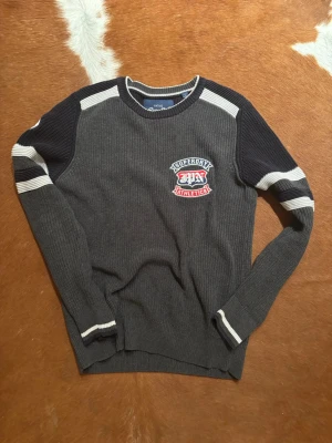 Grå stickad tröja från Superdry - Mörkgrå stickad tröja från Superdry med svarta partier på axlar och ärmar samt vita ränder. Broderad Superdry Athletics-logga på bröstet och ärmen. Rund halsringning och ribbade muddar. Perfekt för en sportig och avslappnad stil.