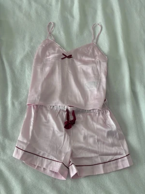 Ljusrosa pyjamasset från Gina Tricot - Söt ljusrosa pyjamasset från Gina Tricot med linne och shorts. Setet har tunna axelband, vinröda rosetter och dekorativa vinröda detaljer längs kanterna. Materialet känns mjukt och lätt mot huden. Fick i present men tyvärr för liten och klippte lappen för snabbt. Endast testad, som ny!